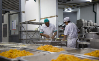 A imagem mostra um ambiente de cozinha industrial ou refeitório. Em primeiro plano, bandejas retangulares cheias de um alimento de cor amarelada, com textura granular, estão dispostas em fileiras, prontas para servir. Ao fundo, duas pessoas vestidos com uniformes de cozinha brancos e toucas (um com touca verde, outro branca) estão trabalhando em uma bancada de aço inoxidável. A pessoa à esquerda manipula o que parece ser um embrulho, enquanto a pessoa à direita está de costas, também manuseando alimentos. Prateleiras metálicas com utensílios de cozinha e recipientes de aço inoxidável são visíveis no fundo à direita e à esquerda.