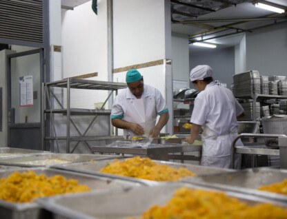 A imagem mostra um ambiente de cozinha industrial ou refeitório. Em primeiro plano, bandejas retangulares cheias de um alimento de cor amarelada, com textura granular, estão dispostas em fileiras, prontas para servir. Ao fundo, duas pessoas vestidos com uniformes de cozinha brancos e toucas (um com touca verde, outro branca) estão trabalhando em uma bancada de aço inoxidável. A pessoa à esquerda manipula o que parece ser um embrulho, enquanto a pessoa à direita está de costas, também manuseando alimentos. Prateleiras metálicas com utensílios de cozinha e recipientes de aço inoxidável são visíveis no fundo à direita e à esquerda.
