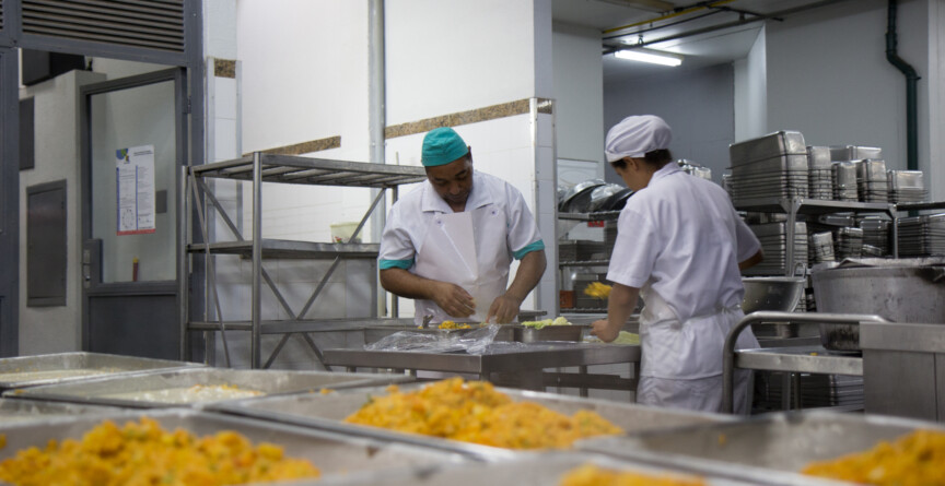A imagem mostra um ambiente de cozinha industrial ou refeitório. Em primeiro plano, bandejas retangulares cheias de um alimento de cor amarelada, com textura granular, estão dispostas em fileiras, prontas para servir. Ao fundo, duas pessoas vestidos com uniformes de cozinha brancos e toucas (um com touca verde, outro branca) estão trabalhando em uma bancada de aço inoxidável. A pessoa à esquerda manipula o que parece ser um embrulho, enquanto a pessoa à direita está de costas, também manuseando alimentos. Prateleiras metálicas com utensílios de cozinha e recipientes de aço inoxidável são visíveis no fundo à direita e à esquerda.