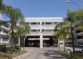 A imagem mostra um grande edifício com uma arquitetura que remete a meados do século XX, com múltiplos andares e uma predominância de concreto e janelas horizontais. Há uma estrutura central que cria uma passagem ou pátio coberto, por onde se pode ver o interior do campus. A área em frente e ao redor do prédio é ajardinada, com palmeiras altas em primeiro plano e outros arbustos. Há um caminho pavimentado que leva à entrada central. Veículos, incluindo carros e motos, estão estacionados na lateral direita da imagem. O céu é azul e claro, indicando um dia ensolarado.