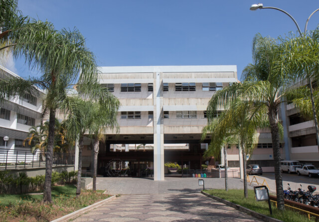 A imagem mostra um grande edifício com uma arquitetura que remete a meados do século XX, com múltiplos andares e uma predominância de concreto e janelas horizontais. Há uma estrutura central que cria uma passagem ou pátio coberto, por onde se pode ver o interior do campus. A área em frente e ao redor do prédio é ajardinada, com palmeiras altas em primeiro plano e outros arbustos. Há um caminho pavimentado que leva à entrada central. Veículos, incluindo carros e motos, estão estacionados na lateral direita da imagem. O céu é azul e claro, indicando um dia ensolarado.