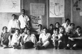 A fotografia em preto e branco registra um grupo de treze jovens, aparentemente estudantes, posando para uma foto em frente a um mural ou parede coberta de cartazes e desenhos. Os jovens estão dispostos em duas fileiras: cinco sentados no chão em primeiro plano e oito em pé ou levemente curvados atrás deles. Os jovens em primeiro plano estão sentados no chão, alguns com as pernas cruzadas ou flexionadas, e apoiam os braços nos joelhos ou no corpo. Na fileira de trás, os jovens estão em pé ou levemente curvados, olhando para a câmera. Suas vestimentas são informais para a época, com camisas de manga curta ou comprida. As expressões faciais variam, com alguns sorrindo e outros com semblante mais sério. A iluminação parece vir de cima e da frente, projetando sombras suaves no chão e na parede. O ambiente sugere um espaço interno, talvez uma sala de aula, um corredor ou um espaço de convivência estudantil.