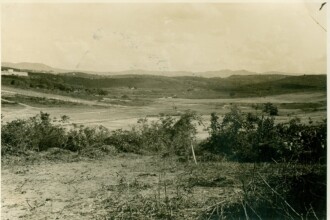 A imagem em preto e branco, com tonalidade sépia, exibe uma paisagem ampla e ondulada de um vale. Em primeiro plano, o terreno é árido e coberto por arbustos baixos e vegetação rasteira, com algumas plantas mais densas à direita. Em segundo plano, o vale se estende com vegetação mais dispersa e algumas áreas planas, que podem ser campos ou terrenos preparados. Ao fundo, uma sequência de colinas e montanhas se eleva, formando o horizonte. No canto superior esquerdo da imagem, é possível avistar um conjunto de edifícios distantes e, em partes da área montanhosa, pequenas manchas que podem indicar construções ou áreas urbanizadas. O céu, na parte superior, é claro e uniforme.
