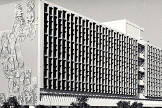 A imagem em preto e branco mostra a fachada de um grande edifício de arquitetura moderna. A parede lateral esquerda apresenta um extenso mural em alto-relevo com figuras humanas estilizadas e elementos abstratos. A parte central e direita do edifício é caracterizada por uma série de janelas verticais e estreitas, dispostas em um padrão repetitivo que cria uma textura visual. No topo do prédio, uma estrutura mais alta e de cor clara se destaca. A base do edifício é ladeada por árvores. O céu, visível ao fundo, é claro e sem nuvens.