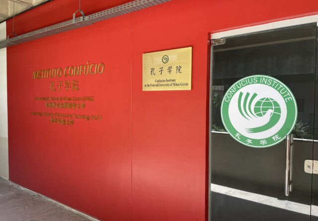 A imagem mostra a entrada de um instituto em um edifício com paredes vermelhas vibrantes. À esquerda, uma placa dourada na parede exibe em relevo "INSTITUTO CONFÚCIO" em português e, abaixo, caracteres chineses. Mais abaixo, há outro texto em português e chinês, de menor tamanho. No centro da parede vermelha, outra placa dourada, menor, traz o texto "孔子学院 Confucius Institute at the Federal University of Minas Gerais". À direita, uma porta de vidro tem um adesivo circular grande e branco com borda verde. Dentro do círculo, há um logotipo em verde que representa uma mão segurando um globo, e também o nome "CONFUCIOUS INSTITUTE" em semicírculo na parte superior e caracteres chineses na parte inferior. O chão é cinza claro, e a iluminação parece ser artificial, vinda de cima.