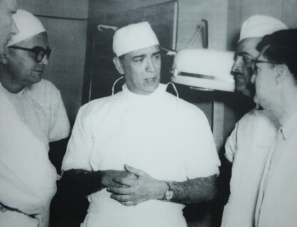 A imagem em preto e branco mostra um grupo de cinco homens em um ambiente que parece ser um centro cirúrgico ou sala de exames. Todos os homens usam uniformes médicos brancos e toucas cirúrgicas brancas. O homem no centro da imagem está de frente para o observador e tem as mãos unidas à frente do corpo, enquanto fala. Um relógio de pulso escuro é visível em seu braço esquerdo. Os outros quatro homens estão ao redor dele, em diferentes posições, aparentemente atentos ao que ele diz. Ao fundo, alguns equipamentos médicos, como uma luminária de teto, podem ser parcialmente vistos.