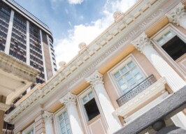 A imagem apresenta a fachada de um edifício histórico em estilo neoclássico, predominantemente na cor pêssego claro com detalhes em branco e dourado. Colunas coríntias decoradas sustentam uma cornija detalhada, sobre a qual está gravado "CONSERVATORIO MINEIRO DE MUSICA" em letras maiúsculas. As janelas retangulares, algumas com sacadas de ferro forjado, estão visíveis. No lado esquerdo da imagem, um arranha-céu moderno, com fachada escura e muitas janelas, eleva-se contrastando com o estilo do Conservatório. O céu acima é azul com algumas nuvens brancas visíveis. O ponto de vista da foto é de baixo para cima, enfatizando a grandiosidade dos edifícios