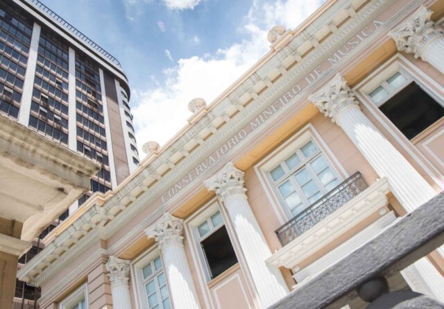 A imagem apresenta a fachada de um edifício histórico em estilo neoclássico, predominantemente na cor pêssego claro com detalhes em branco e dourado. Colunas coríntias decoradas sustentam uma cornija detalhada, sobre a qual está gravado "CONSERVATORIO MINEIRO DE MUSICA" em letras maiúsculas. As janelas retangulares, algumas com sacadas de ferro forjado, estão visíveis. No lado esquerdo da imagem, um arranha-céu moderno, com fachada escura e muitas janelas, eleva-se contrastando com o estilo do Conservatório. O céu acima é azul com algumas nuvens brancas visíveis. O ponto de vista da foto é de baixo para cima, enfatizando a grandiosidade dos edifícios