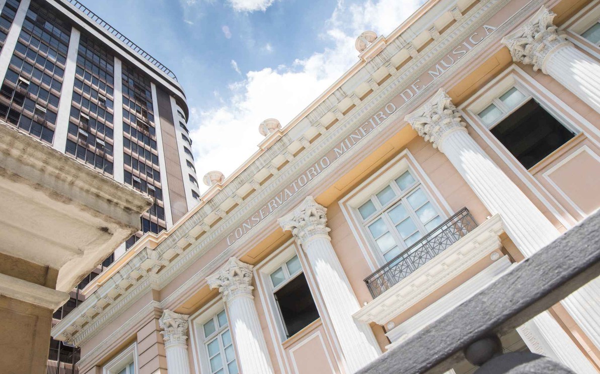 A imagem apresenta a fachada de um edifício histórico em estilo neoclássico, predominantemente na cor pêssego claro com detalhes em branco e dourado. Colunas coríntias decoradas sustentam uma cornija detalhada, sobre a qual está gravado "CONSERVATORIO MINEIRO DE MUSICA" em letras maiúsculas. As janelas retangulares, algumas com sacadas de ferro forjado, estão visíveis. No lado esquerdo da imagem, um arranha-céu moderno, com fachada escura e muitas janelas, eleva-se contrastando com o estilo do Conservatório. O céu acima é azul com algumas nuvens brancas visíveis. O ponto de vista da foto é de baixo para cima, enfatizando a grandiosidade dos edifícios