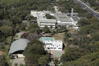 A imagem é uma fotografia aérea de uma área arborizada, que abriga diversos edifícios e instalações esportivas. No centro, um grande complexo de prédios brancos de dois andares com telhados planos se destaca, cercado por uma densa vegetação. As janelas dos edifícios são retangulares e há uma torre de água ou chaminé no telhado. À frente do complexo principal, um prédio branco de formato alongado com janelas circulares está localizado próximo a uma piscina retangular de água azul. Ao lado da piscina, há duas quadras de tênis de saibro. Na parte inferior esquerda da imagem, um ginásio com um telhado de metal arqueado é visível. Uma estrada com tráfego de veículos percorre a parte superior direita da foto.
