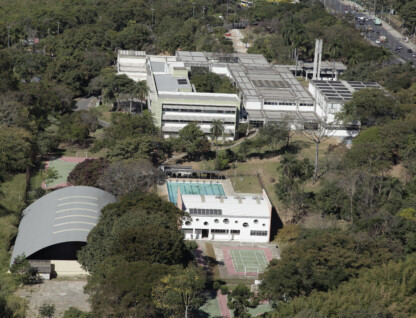 A imagem é uma fotografia aérea de uma área arborizada, que abriga diversos edifícios e instalações esportivas. No centro, um grande complexo de prédios brancos de dois andares com telhados planos se destaca, cercado por uma densa vegetação. As janelas dos edifícios são retangulares e há uma torre de água ou chaminé no telhado. À frente do complexo principal, um prédio branco de formato alongado com janelas circulares está localizado próximo a uma piscina retangular de água azul. Ao lado da piscina, há duas quadras de tênis de saibro. Na parte inferior esquerda da imagem, um ginásio com um telhado de metal arqueado é visível. Uma estrada com tráfego de veículos percorre a parte superior direita da foto.