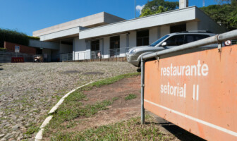 A imagem mostra um edifício de fachada branca, com um telhado de dois andares, sob um céu azul e com nuvens brancas. Na frente do edifício, uma placa de cor laranja com letras brancas informa "restaurante setorial II". O edifício tem uma rampa de acesso, portas de vidro e janelas retangulares. Um carro cinza está estacionado à direita, e a rua, em primeiro plano, é feita de paralelepípedos.