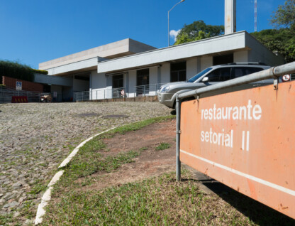A imagem mostra um edifício de fachada branca, com um telhado de dois andares, sob um céu azul e com nuvens brancas. Na frente do edifício, uma placa de cor laranja com letras brancas informa "restaurante setorial II". O edifício tem uma rampa de acesso, portas de vidro e janelas retangulares. Um carro cinza está estacionado à direita, e a rua, em primeiro plano, é feita de paralelepípedos.