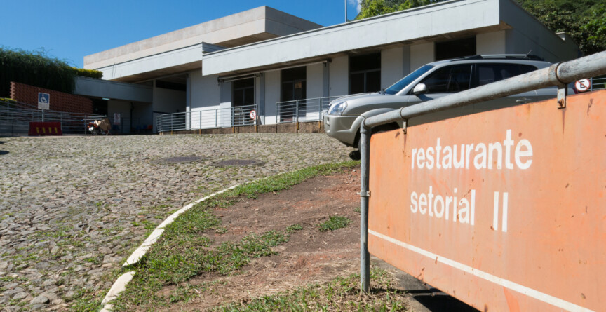 A imagem mostra um edifício de fachada branca, com um telhado de dois andares, sob um céu azul e com nuvens brancas. Na frente do edifício, uma placa de cor laranja com letras brancas informa "restaurante setorial II". O edifício tem uma rampa de acesso, portas de vidro e janelas retangulares. Um carro cinza está estacionado à direita, e a rua, em primeiro plano, é feita de paralelepípedos.