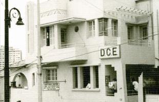 A imagem em preto e branco mostra um edifício de três andares com arquitetura Art Déco. A fachada do prédio é composta por linhas retas, janelas com grades e uma varanda no andar superior. No centro da fachada, há um letreiro com as letras "DCE". À esquerda, uma luminária de rua antiga com detalhes em metal é visível. Ao fundo, um edifício mais alto, provavelmente um arranha-céu, é parcialmente visível, enquanto fios de eletricidade cortam a imagem.