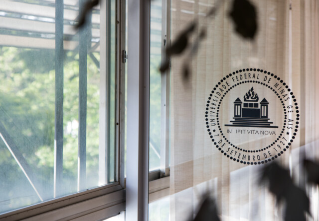 Essa imagem mostra uma janela, com um selo ou adesivo da Universidade Federal de Minas Gerais (UFMG) visível na parte direita do vidro. O selo circular mostra um edifício com uma chama no topo e a inscrição "UNIVERSIDADE FEDERAL DE MINAS GERAIS" e a data "7 DE SETEMBRO DE 1927" ao redor, juntamente com o lema "INCIPIT VITA NOVA". O sol e a vegetação do lado de fora criam reflexos e um brilho suave no vidro da janela. A imagem tem profundidade, com o selo em foco nítido no primeiro plano, enquanto a paisagem externa — incluindo grades e a folhagem de árvores — aparece desfocada ao fundo. A iluminação geral é clara e natural.
