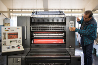 A imagem mostra um homem com uma jaqueta xadrez azul e jeans, em pé ao lado de uma grande máquina de impressão em um ambiente interno. Ele está segurando a alça da máquina com as duas mãos e olhando para os botões e controles na lateral. A máquina é de cor cinza e preta, com um cilindro vermelho visível no centro. Um painel de controle branco com luzes e botões está à esquerda. O ambiente, que parece ser uma oficina ou laboratório, tem uma janela com grades ao fundo e lâmpadas penduradas no teto.