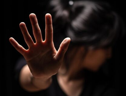 Mulher com a mão levantada em direção à câmera, em um gesto de "pare", com o rosto virado e desfocado ao fundo