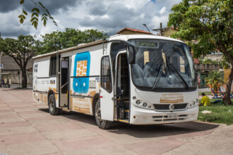 A imagem mostra um ônibus branco com detalhes em azul e amarelo, com a inscrição "Carro-Biblioteca e Telecentro UFMG" nas laterais. O ônibus está estacionado em uma praça com piso de paralelepípedos e árvores ao redor. A porta do ônibus está aberta, e a escadaria é visível, enquanto o vidro dianteiro tem a inscrição "ESPECIAL". O fundo da imagem revela um céu nublado, com um prédio e árvores.