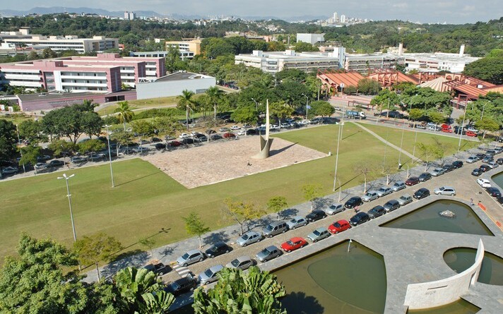 A imagem é uma vista aérea de um campus universitário com áreas verdes, edifícios e estacionamentos. No centro da imagem, há um grande gramado com um monumento de metal no centro, cercado por um caminho pavimentado. À esquerda e à direita do gramado, há longas fileiras de carros estacionados em vagas ao ar livre. Ao fundo, uma série de edifícios de diferentes cores e tamanhos se estendem, incluindo um prédio cor-de-rosa no lado esquerdo. Árvores e vegetação preenchem as áreas entre os edifícios. Na parte inferior direita, um espelho d'água de formato irregular é visível. Ao longe, uma paisagem de cidade é vista no horizonte.