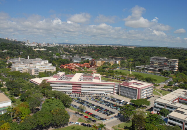 The image shows an elevated, wide-angle shot of a sprawling university campus on a sunny day. In the foreground, a large, modern building with a red and white facade dominates the view, with a massive parking lot full of cars directly in front of it. The campus is very green, with many trees and patches of forest interspersed between the various academic buildings.  Beyond the central building, more campus facilities can be seen, including a sports field and several other multi-story structures of varying architectural styles. A city skyline is visible in the distance on the left side of the frame, suggesting the university is located in or on the outskirts of a major metropolitan area. The sky is bright blue with scattered white clouds. The overall impression is one of a large, well-established institution set within a lush, hilly landscape.