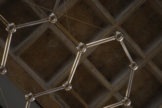 A imagem mostra uma estrutura suspensa em um teto de concreto. A estrutura é composta por tubos metálicos prateados, interligados por esferas brilhantes, formando um padrão hexagonal. Vários fios finos de metal, quase imperceptíveis, prendem a estrutura ao teto. O teto é de concreto, com uma textura de painéis quadrados em formato de favo de mel. A iluminação ambiente é difusa e as superfícies metálicas da estrutura refletem a luz. A foto é tirada de um ângulo que mostra a estrutura de baixo para cima.