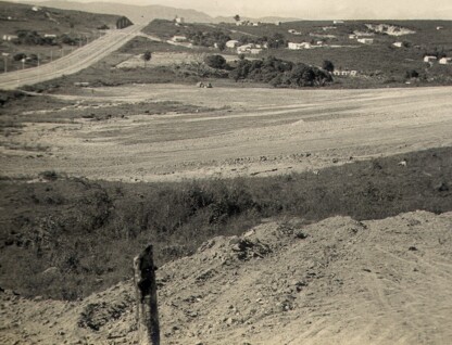 A imagem em preto e branco mostra uma paisagem de terreno aberto e em declive. A cena é dominada por solo exposto e áreas de vegetação rasteira. Em primeiro plano, há um monte de terra com um toco de madeira. No plano intermediário, há um trecho de estrada de terra visível e, mais ao fundo, a topografia se eleva com casas esparsas e vegetação. Do lado esquerdo, no topo, uma estrada pavimentada com postes de luz segue a curva da colina, desaparecendo no horizonte.