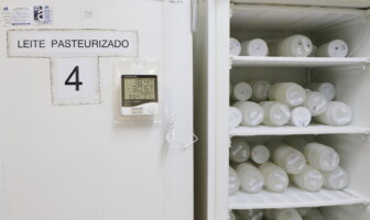 imagem mostra um refrigerador branco em um ambiente interno. A porta do refrigerador à esquerda tem uma placa com o texto "LEITE PASTEURIZADO" e o número "4". Um termômetro digital está fixado na porta, exibindo as temperaturas "18.4°C" e "-30.5°C". A porta do refrigerador está aberta, revelando seu interior com várias prateleiras. As prateleiras estão cheias de potes ou frascos de plástico, brancos e transparentes, dispostos lado a lado. A iluminação é difusa, e o ambiente parece ser um laboratório ou um espaço de armazenamento.