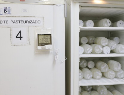 imagem mostra um refrigerador branco em um ambiente interno. A porta do refrigerador à esquerda tem uma placa com o texto "LEITE PASTEURIZADO" e o número "4". Um termômetro digital está fixado na porta, exibindo as temperaturas "18.4°C" e "-30.5°C". A porta do refrigerador está aberta, revelando seu interior com várias prateleiras. As prateleiras estão cheias de potes ou frascos de plástico, brancos e transparentes, dispostos lado a lado. A iluminação é difusa, e o ambiente parece ser um laboratório ou um espaço de armazenamento.