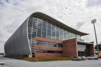 A imagem mostra a vista externa de um ginásio moderno com uma arquitetura arrojada. O edifício, que ocupa a maior parte da imagem, tem a fachada de tijolos aparentes na parte inferior e uma estrutura curva com painéis de metal cinza na parte superior. Grandes janelas de vidro compõem a maior parte da parede do prédio, permitindo que a luz do sol entre. Na frente do prédio, a área é coberta por brita cinza, e três carros, dois brancos e um cinza, estão estacionados. À direita, um poste alto de iluminação de quadra esportiva é visível. O céu é parcialmente nublado, com nuvens brancas e cinzas.