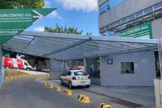 A imagem mostra a entrada de um edifício, a Unidade de Pronto Atendimento (UPA) Centro-Sul. A entrada tem uma cobertura de vidro e metal. Acima, uma grande placa triangular verde com letras brancas indica "UPA CENTRO-SUL", "UNIDADE DE PRONTO ATENDIMENTO", e "DA PREFEITURA DE BELO HORIZONTE". Há outra placa similar no prédio. Em primeiro plano, uma via de acesso é visível, com blocos amarelos de concreto no chão. Um carro branco com o emblema da "Guarda Civil Municipal" está estacionado na área de acesso. Ao fundo, uma ambulância branca e vermelha e outros veículos estão estacionados. O edifício tem paredes cinzas, e algumas janelas e placas de sinalização são visíveis. O céu está azul, com nuvens brancas.