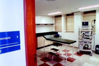 A imagem mostra o interior de uma sala de exames em um ambiente clínico. O piso é coberto por azulejos quadrados, alternando entre cores claras e escuras, formando um padrão xadrez. À esquerda, uma porta de madeira parcialmente aberta exibe uma placa azul e branca com os dizeres "Endoscopia", "2" e "212". No centro da sala, há uma maca de exame com estofamento escuro e uma escada de dois degraus ao lado. Ao fundo, a sala tem paredes brancas e janelas com persianas de madeira. Um armário de cor clara está fixado na parede. À direita da maca, um carrinho de metal com rodas abriga vários equipamentos eletrônicos e monitores, típicos de exames médicos. Um malote de cor clara com a logomarca de uma empresa está no chão, ao lado do carrinho. A sala é iluminada por luzes fluorescentes no teto.