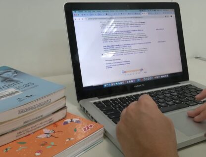 Imagem mostra as maos de uma pessoa mexendo em um computador com livros ao lado