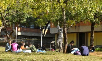 A imagem mostra um grupo de pessoas sentadas na grama, em um ambiente externo, à sombra de árvores. No primeiro plano, à esquerda, quatro pessoas estão sentadas em círculo, conversando e lendo. Outras pessoas estão sentadas individualmente ou em pequenos grupos. A pessoa no canto inferior esquerdo está de camisa rosa, sentada de frente para as outras. Entre as árvores, uma estátua escura de uma figura feminina está visível. O chão é coberto por grama verde, com algumas folhas secas. Ao fundo, um prédio de tijolos vermelhos e grandes janelas retangulares é visível. A luz do sol é intensa e cria áreas de sombra.