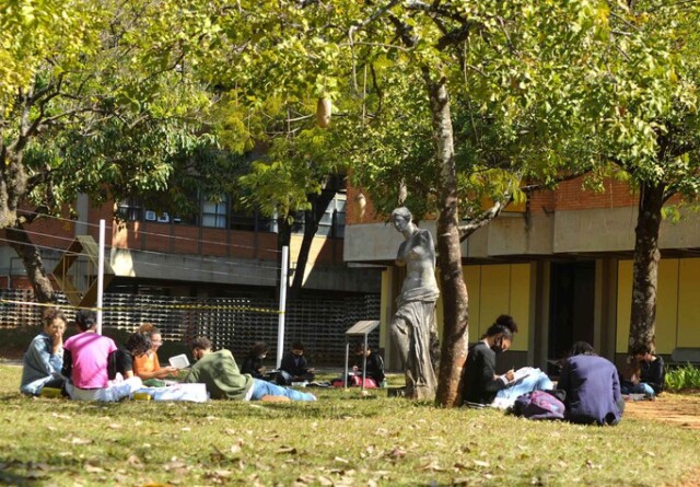 A imagem mostra um grupo de pessoas sentadas na grama, em um ambiente externo, à sombra de árvores. No primeiro plano, à esquerda, quatro pessoas estão sentadas em círculo, conversando e lendo. Outras pessoas estão sentadas individualmente ou em pequenos grupos. A pessoa no canto inferior esquerdo está de camisa rosa, sentada de frente para as outras. Entre as árvores, uma estátua escura de uma figura feminina está visível. O chão é coberto por grama verde, com algumas folhas secas. Ao fundo, um prédio de tijolos vermelhos e grandes janelas retangulares é visível. A luz do sol é intensa e cria áreas de sombra.
