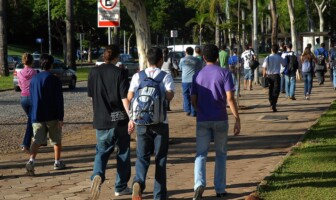 A imagem mostra um grupo de pessoas caminhando em uma calçada de blocos de cimento em um ambiente externo. No primeiro plano, três jovens, vistos de costas, estão caminhando em direção ao centro da imagem. O do meio usa uma mochila azul e branca. Ao fundo, há um estacionamento com carros, cercado por grama e árvores. Outras pessoas, em grupos e sozinhas, estão caminhando na mesma direção. Uma placa de trânsito branca, com o símbolo de "E" cortado, indicando proibido estacionar, está visível no canto superior esquerdo. A iluminação é natural, proveniente da luz do sol.