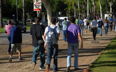 A imagem mostra um grupo de pessoas caminhando em uma calçada de blocos de cimento em um ambiente externo. No primeiro plano, três jovens, vistos de costas, estão caminhando em direção ao centro da imagem. O do meio usa uma mochila azul e branca. Ao fundo, há um estacionamento com carros, cercado por grama e árvores. Outras pessoas, em grupos e sozinhas, estão caminhando na mesma direção. Uma placa de trânsito branca, com o símbolo de "E" cortado, indicando proibido estacionar, está visível no canto superior esquerdo. A iluminação é natural, proveniente da luz do sol.