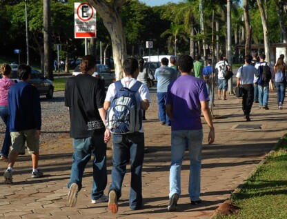 A imagem mostra um grupo de pessoas caminhando em uma calçada de blocos de cimento em um ambiente externo. No primeiro plano, três jovens, vistos de costas, estão caminhando em direção ao centro da imagem. O do meio usa uma mochila azul e branca. Ao fundo, há um estacionamento com carros, cercado por grama e árvores. Outras pessoas, em grupos e sozinhas, estão caminhando na mesma direção. Uma placa de trânsito branca, com o símbolo de "E" cortado, indicando proibido estacionar, está visível no canto superior esquerdo. A iluminação é natural, proveniente da luz do sol.