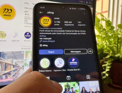 Imagem mostra, em primeiro plano, mão de pessoa segurando o celular, com o Instagram da UFMG aberto, e, em segundo plano, mesmo Instagram sendo mostrado em um monitor