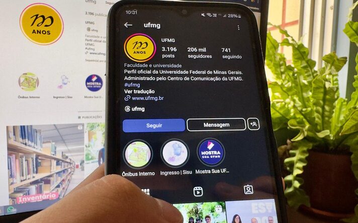 Imagem mostra, em primeiro plano, mão de pessoa segurando o celular, com o Instagram da UFMG aberto, e, em segundo plano, mesmo Instagram sendo mostrado em um monitor