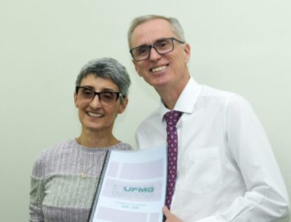 Alamanda é uma mulher branca, usa óculos e veste uma blusa cinza; Alessandro é um homem branco, de camisa branca e gravata; ele segura um documento nas mãos