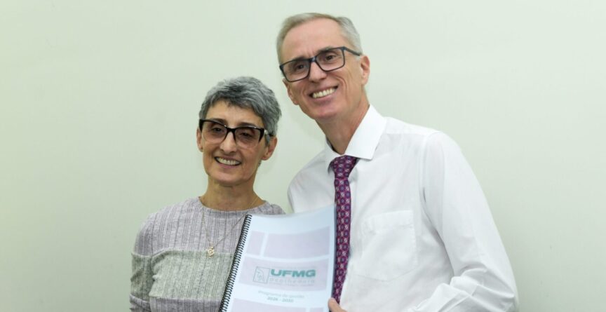 Alamanda é uma mulher branca, usa óculos e veste uma blusa cinza; Alessandro é um homem branco, de camisa branca e gravata; ele segura um documento nas mãos