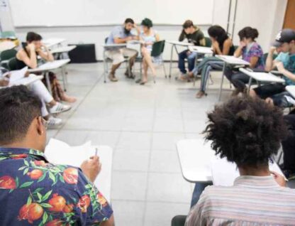 Imagem mostra sala com diversas pessoas sentadas em carteiras escolares, dispostas em forma de um círculo