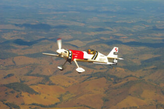 A imagem mostra um avião de acrobacia voando em um céu azul com nuvens brancas, sobre uma paisagem de colinas e campos. O avião é branco e tem uma cauda vermelha, uma faixa vermelha na parte superior da fuselagem e logotipos em preto e amarelo. Ele tem asas curtas e uma única hélice, e a cabine é visível. A paisagem abaixo é montanhosa e coberta por vegetação de cor marrom e verde. É possível ver algumas estradas de terra no meio das colinas. A imagem é vista de uma perspectiva elevada, mostrando a grande extensão das montanhas e campos.
