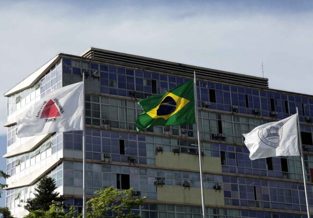A imagem mostra a fachada de um edifício alto e moderno, com uma arquitetura de vidro e concreto, refletindo o céu. Em frente ao prédio, três mastros exibem bandeiras que tremulam ao vento. Da esquerda para a direita, as bandeiras são: A bandeira do estado de Minas Gerais, branca com um triângulo vermelho no centro e a inscrição "Libertas Quae Sera Tamen". instituição, com suas cores verde, amarela, azul e branca, destacando-se no centro da composição. A bandeira da Universidade Federal de Minas Gerais, branca com o brasão da universidade ao centro. O céu, com algumas nuvens dispersas, serve de pano de fundo para as bandeiras e o edifício, sugerindo um dia claro e com vento. A foto transmite uma atmosfera de representação oficial e institucional.