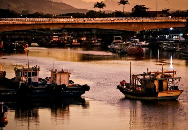Imagem ilustra Pontal da Barra de Macaé, que integra a Bacia de Campos, com alguns barcos em primeiro plano e, ao fundo, outras embarcações e uma ponte, no final de tarde