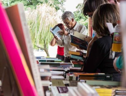Imagem mostra livro em primeiro plano e, ao fundo, pessoas vendo outros livros em uma feira