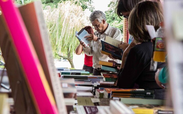 Imagem mostra livro em primeiro plano e, ao fundo, pessoas vendo outros livros em uma feira