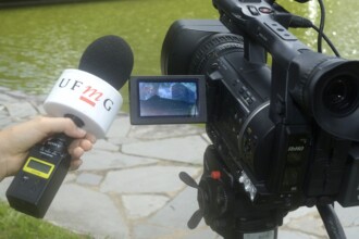 A imagem mostra uma mão segurando um microfone de entrevista em primeiro plano, com uma câmera de vídeo profissional ao lado. O microfone tem um protetor de espuma preto, e uma "cubo" branco com as letras "UFMG" em vermelho. A mão segura o microfone pela haste, conectada a um equipamento transmissor. A câmera de vídeo é preta e grande, com uma tela pequena, que mostra a imagem de um edifício. A câmera está em um tripé. No fundo, há um corpo d'água com a superfície verde-escura, cercado por um caminho pavimentado de pedras.