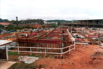 A imagem mostra uma vista panorâmica de um canteiro de obras em um terreno de terra vermelha. Em primeiro plano e no centro, uma estrutura de concreto em construção, com paredes e colunas de cimento reforçado com barras de metal, forma o que parece ser um fosso. Andaimes de madeira e cercas de proteção brancas de metal estão ao redor da estrutura, e dois trabalhadores podem ser vistos no local. Vários materiais de construção, como tábuas e vergalhões, estão espalhados pelo chão. Ao fundo, à direita, há outro prédio em construção, com uma estrutura de vários andares visível. À esquerda, uma rua pavimentada é visível com carros estacionados ao longo dela e algumas pessoas caminhando na calçada. A paisagem é composta por árvores, e, ao longe, outras construções e prédios podem ser observados. O céu está coberto de nuvens, em um dia claro, e as sombras são pouco evidentes.